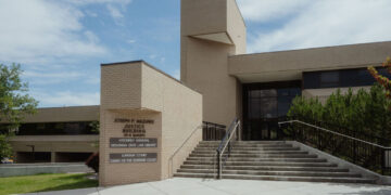 Exterior view of the Joseph P. Mazurek Justice Building with wide concrete steps and railing, beige brick facade, and a prominent sign in the foreground.