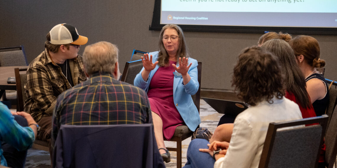 Woman in a blue blazer speaks to a small audience at a panel, with a bulleted slide projected behind her.