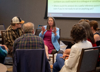 Woman in a blue blazer speaks to a small audience at a panel, with a bulleted slide projected behind her.