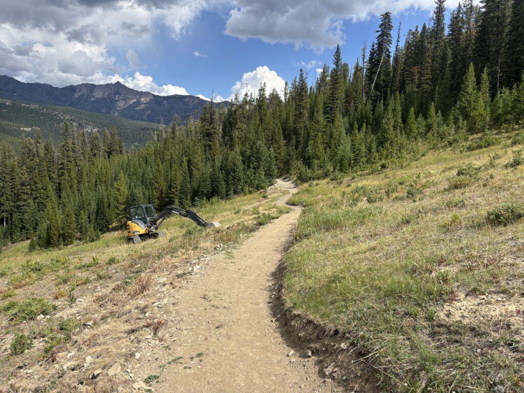 Dirt hiking trail winds through a forested hillside with mountains in the distance and cloudy sky above, with a yellow excavator partially visible on the lower left.