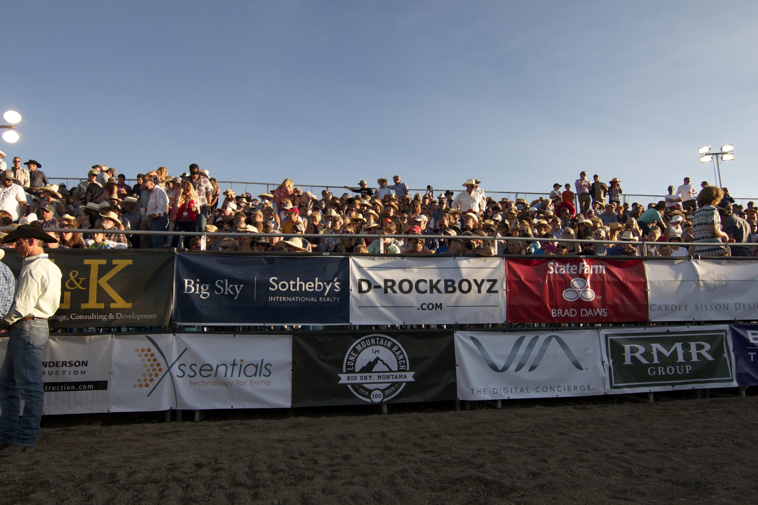 Crowd of spectators seated in stadium bleachers behind sponsor banners at an outdoor event against a blue sky.