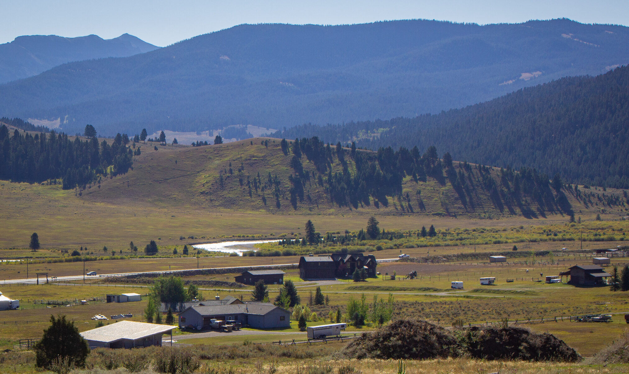 Rural valley with farm buildings at the base of tree-covered hills and distant blue mountains under a clear sky