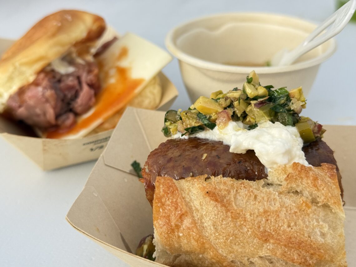 Close-up of a crusty bun sandwich topped with a white spread and chunky relish; a cup and spoon are blurred in the background.