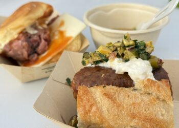 Close-up of a crusty bun sandwich topped with a white spread and chunky relish; a cup and spoon are blurred in the background.