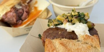 Close-up of a crusty bun sandwich topped with a white spread and chunky relish; a cup and spoon are blurred in the background.
