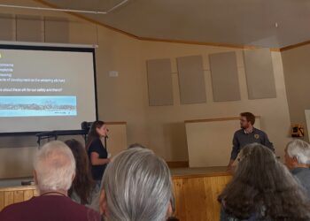 Presentation in a meeting hall: slide titled 'Study Area' with bullets on community concerns; two presenters stand near screen as audience listens.