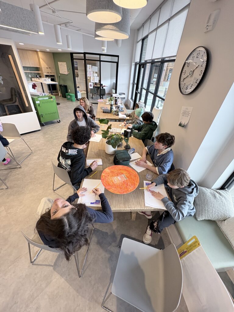 Group of students gathered around a long wooden table, drawing with pencils in a bright, modern classroom with large windows and a wall clock visible.