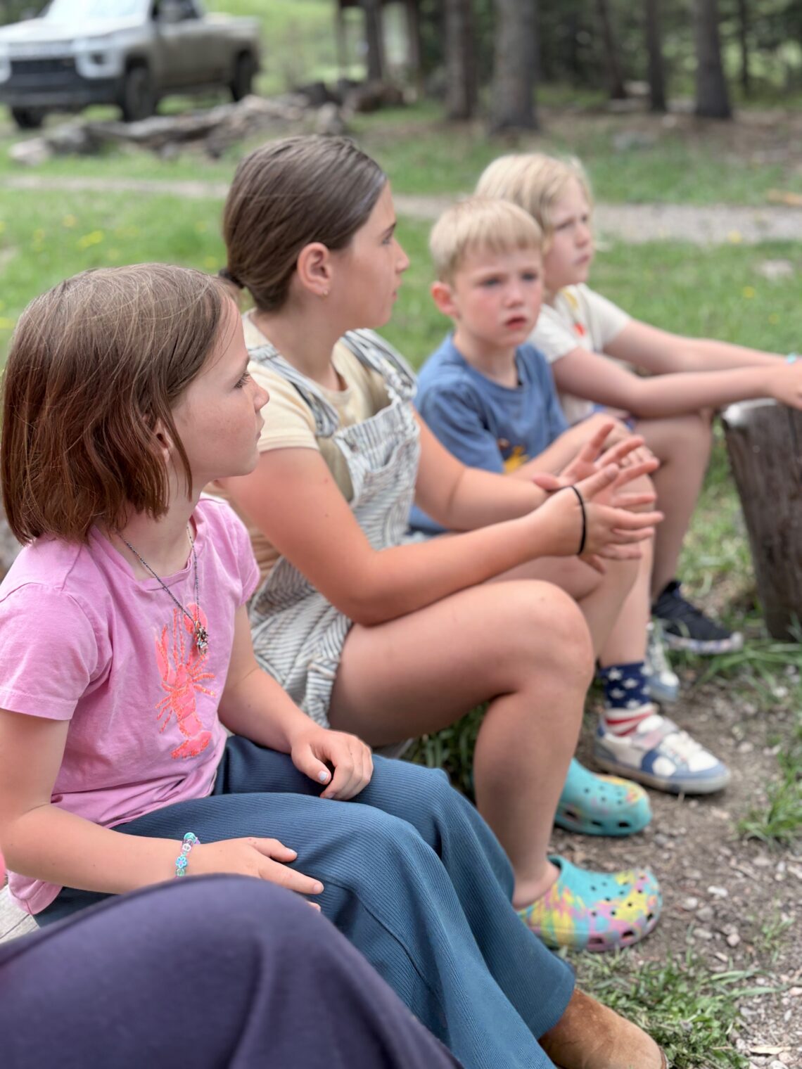 Four children sit on a wooden log in a grassy park, hands folded, wearing casual clothes.