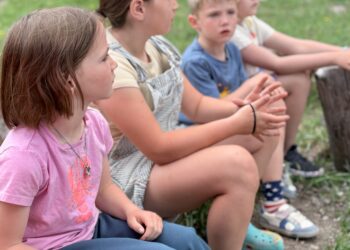 Four children sit on a wooden log in a grassy park, hands folded, wearing casual clothes.