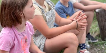 Four children sit on a wooden log in a grassy park, hands folded, wearing casual clothes.
