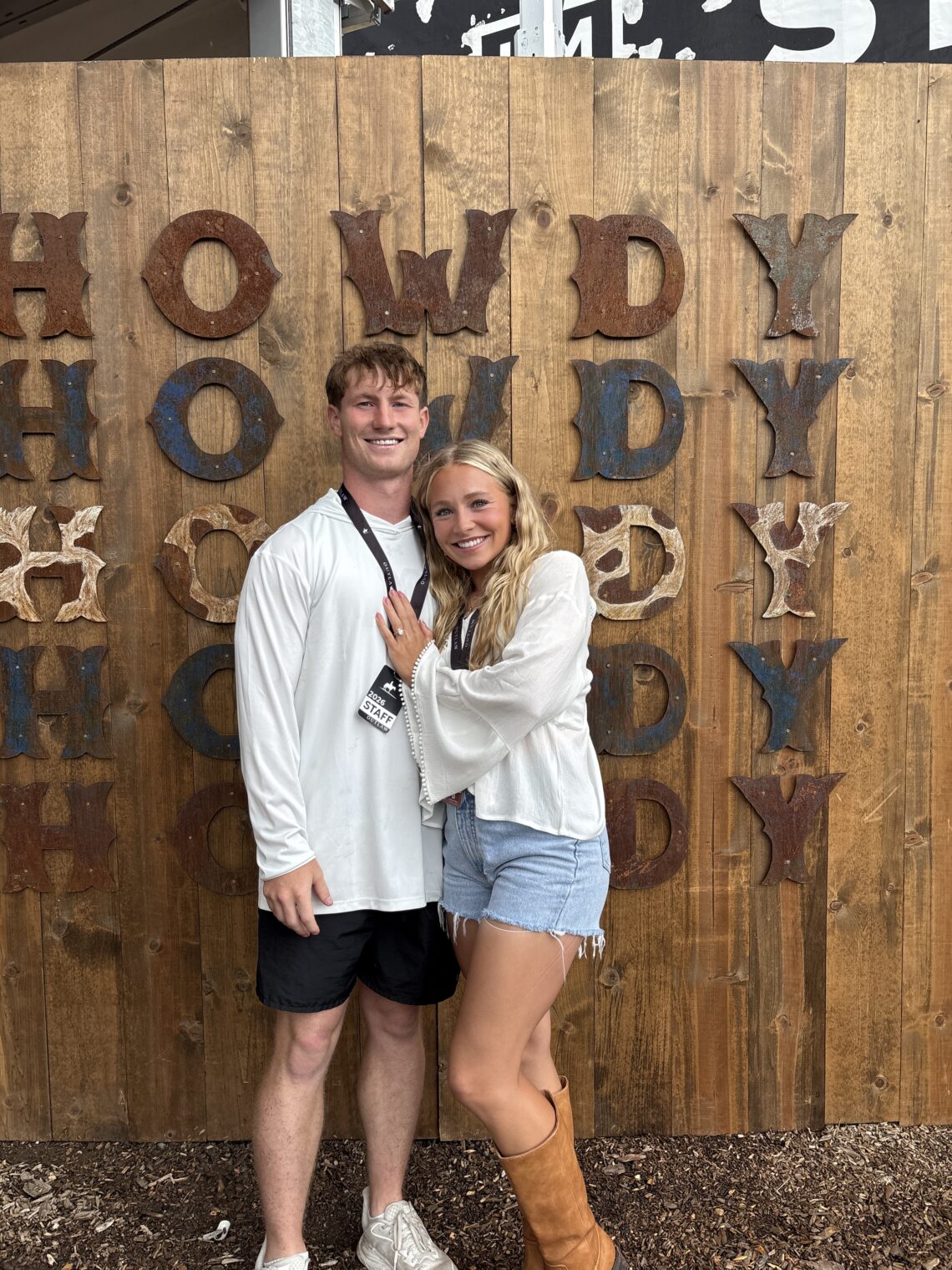 Smiling couple standing arm in arm in front of a rustic wooden wall with large rusted letters behind them.