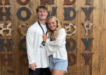 Smiling couple standing arm in arm in front of a rustic wooden wall with large rusted letters behind them.