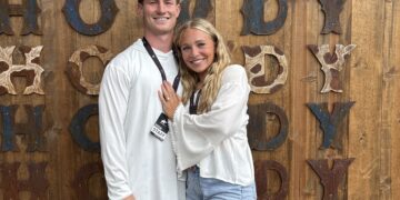 Smiling couple standing arm in arm in front of a rustic wooden wall with large rusted letters behind them.