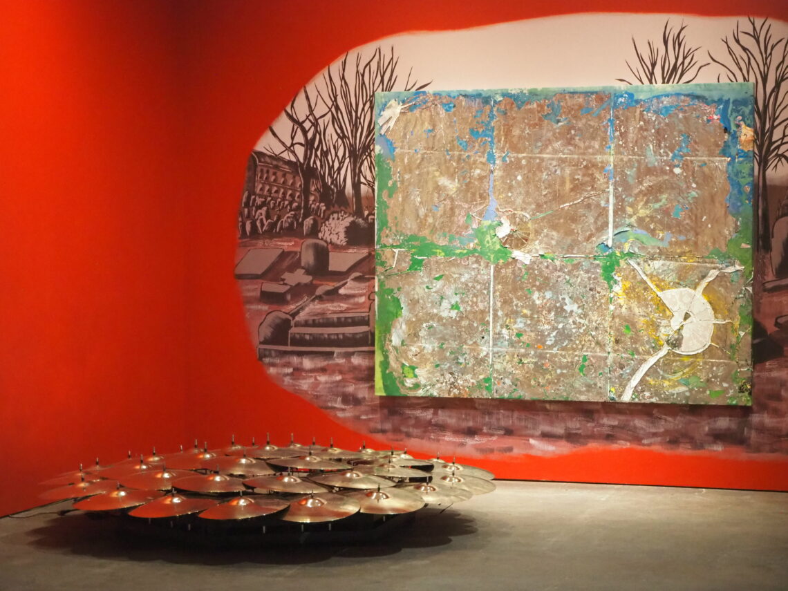 A drum kit with many cymbals on a stage, set against a bright red curved wall and a large abstract mural behind it.