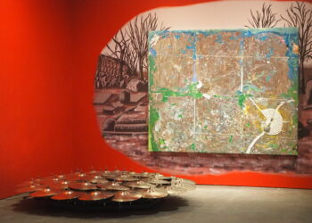 A drum kit with many cymbals on a stage, set against a bright red curved wall and a large abstract mural behind it.