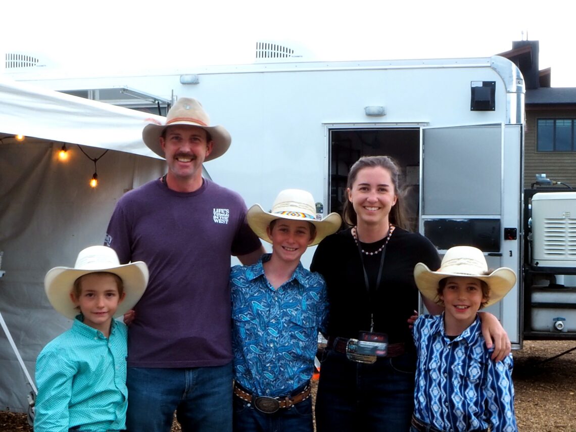 Group of five people in cowboy hats posing in front of a white trailer, with string lights visible on the left.