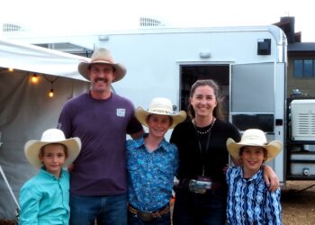 Group of five people in cowboy hats posing in front of a white trailer, with string lights visible on the left.