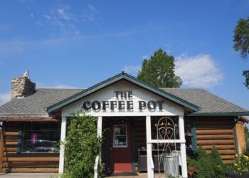Front view of a rustic coffee shop with a peaked roof that reads 'The Coffee Pot' on the facade and a stone chimney on the left.