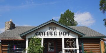 Front view of a rustic coffee shop with a peaked roof that reads 'The Coffee Pot' on the facade and a stone chimney on the left.