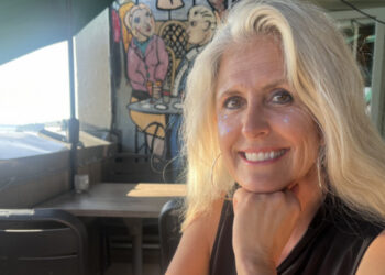Smiling blonde woman resting chin on her hand at an outdoor cafe, with colorful mural behind her and glitter on her cheeks.