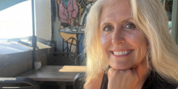 Smiling blonde woman resting chin on her hand at an outdoor cafe, with colorful mural behind her and glitter on her cheeks.