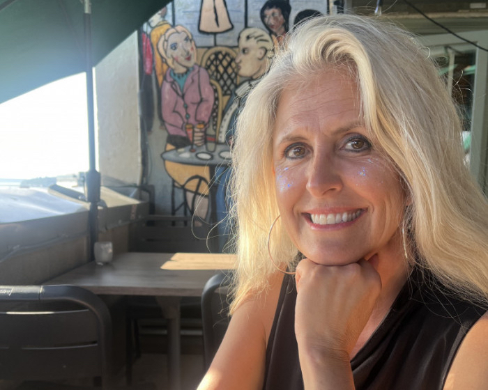 Smiling blonde woman resting chin on her hand at an outdoor cafe, with colorful mural behind her and glitter on her cheeks.