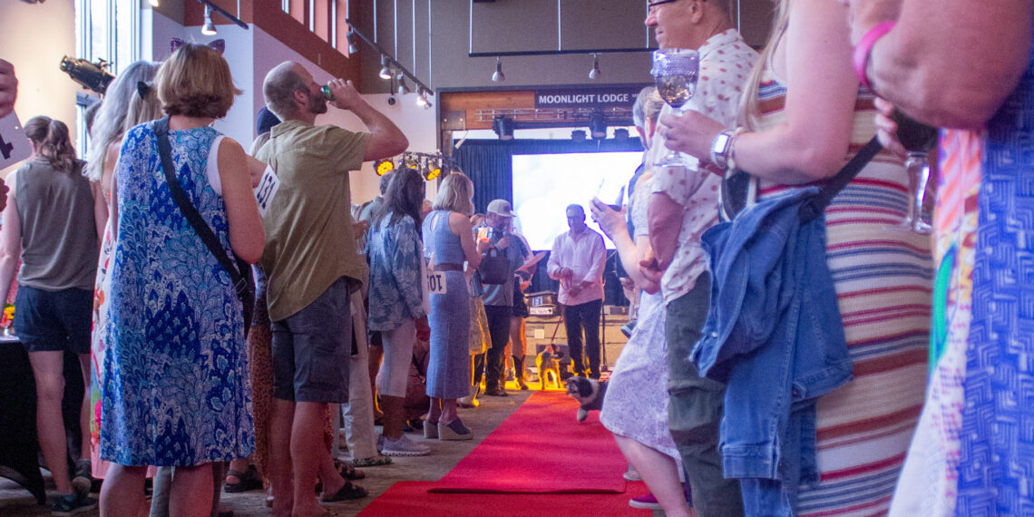 People lined up along a red carpet indoors, some holding drinks, with a 'Moonlight Lodge' sign in the background.