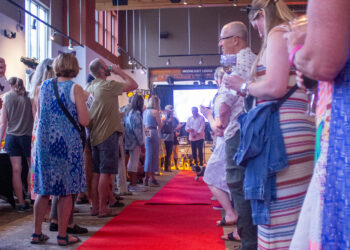 People lined up along a red carpet indoors, some holding drinks, with a 'Moonlight Lodge' sign in the background.