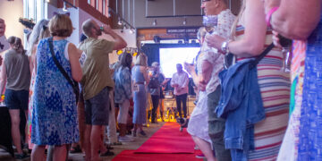 People lined up along a red carpet indoors, some holding drinks, with a 'Moonlight Lodge' sign in the background.