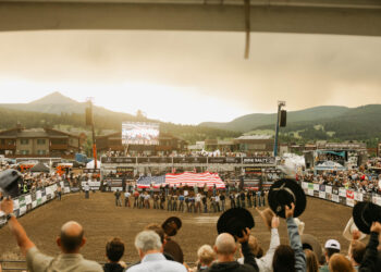 Outdoor arena filled with spectators as a large American flag is displayed over the ring, with banners along the rail and mountains in the background at sunset.