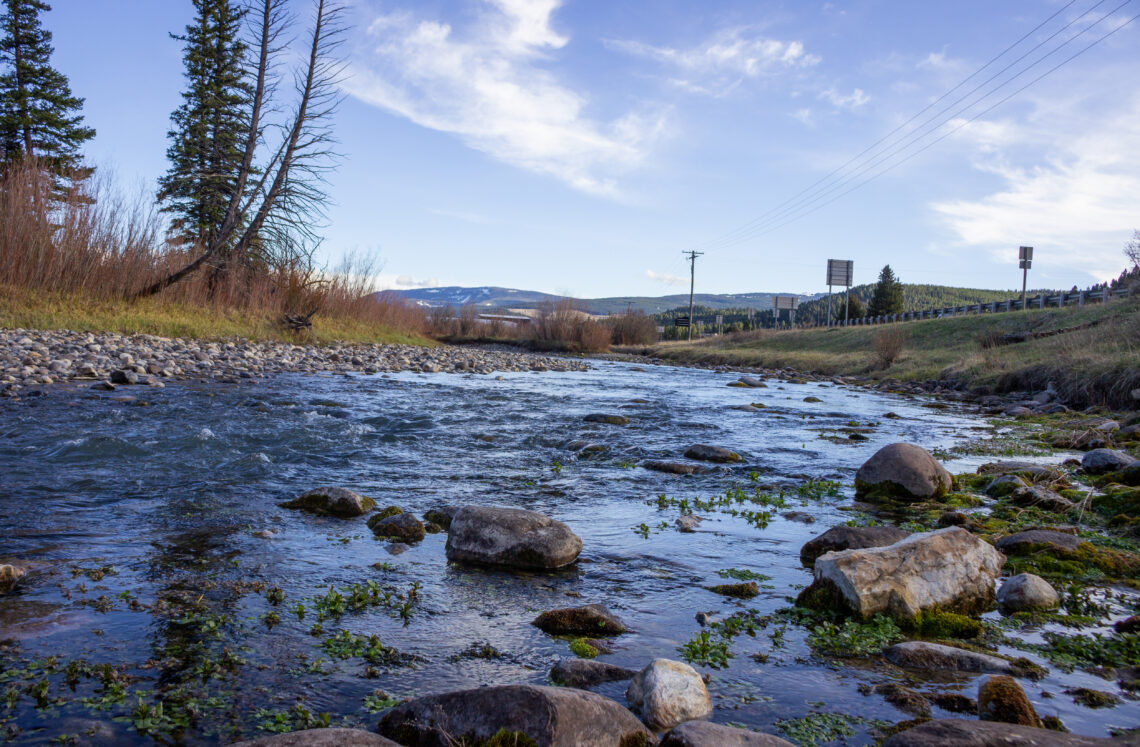 Shallow river with large rocks along the bank, bare shrubs, and distant hills under a blue sky with a few clouds.