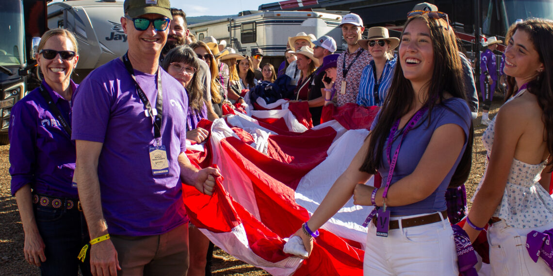 Smiling group of volunteers in purple shirts and lanyards standing outdoors with a large red-and-white banner.