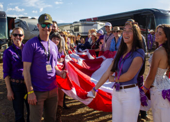Smiling group of volunteers in purple shirts and lanyards standing outdoors with a large red-and-white banner.