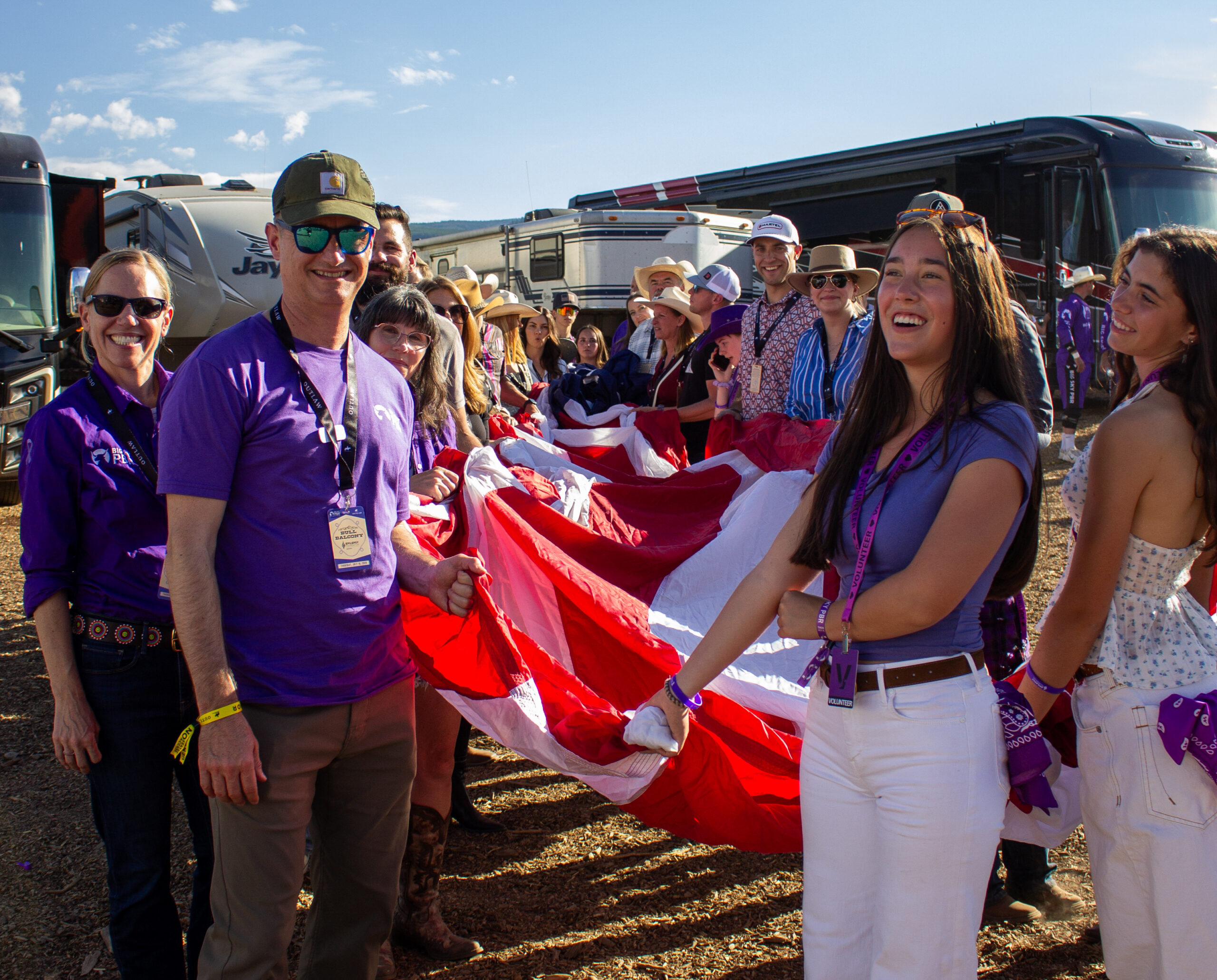 Smiling group of volunteers in purple shirts and lanyards standing outdoors with a large red-and-white banner.