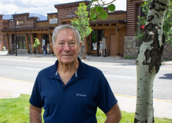Portrait of an older man in a navy Columbia polo standing on a sidewalk with wooden storefronts and a tree behind him.