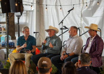 Four men seated on a stage in front of an audience, each with a microphone, wearing casual shirts and cowboy hats during a panel discussion under string lights inside a tent.