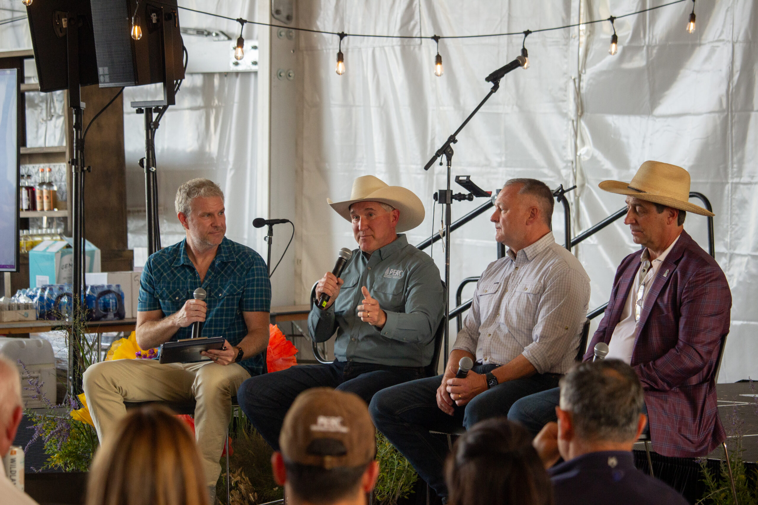Four men seated on a stage in front of an audience, each with a microphone, wearing casual shirts and cowboy hats during a panel discussion under string lights inside a tent.