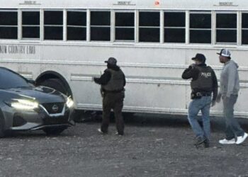 White school bus parked on a gravel lot with two officers and a man in a gray hoodie nearby; a silver car is in the foreground, suggesting a stop or encounter.