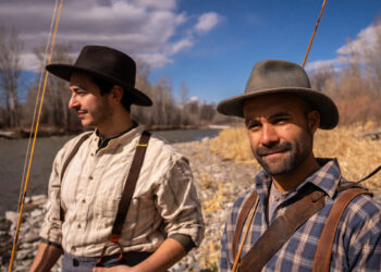 Two men in vintage-style outfits fishing by a rocky riverbank on a sunny day, wearing wide-brim hats and suspenders.
