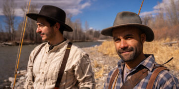 Two men in vintage-style outfits fishing by a rocky riverbank on a sunny day, wearing wide-brim hats and suspenders.