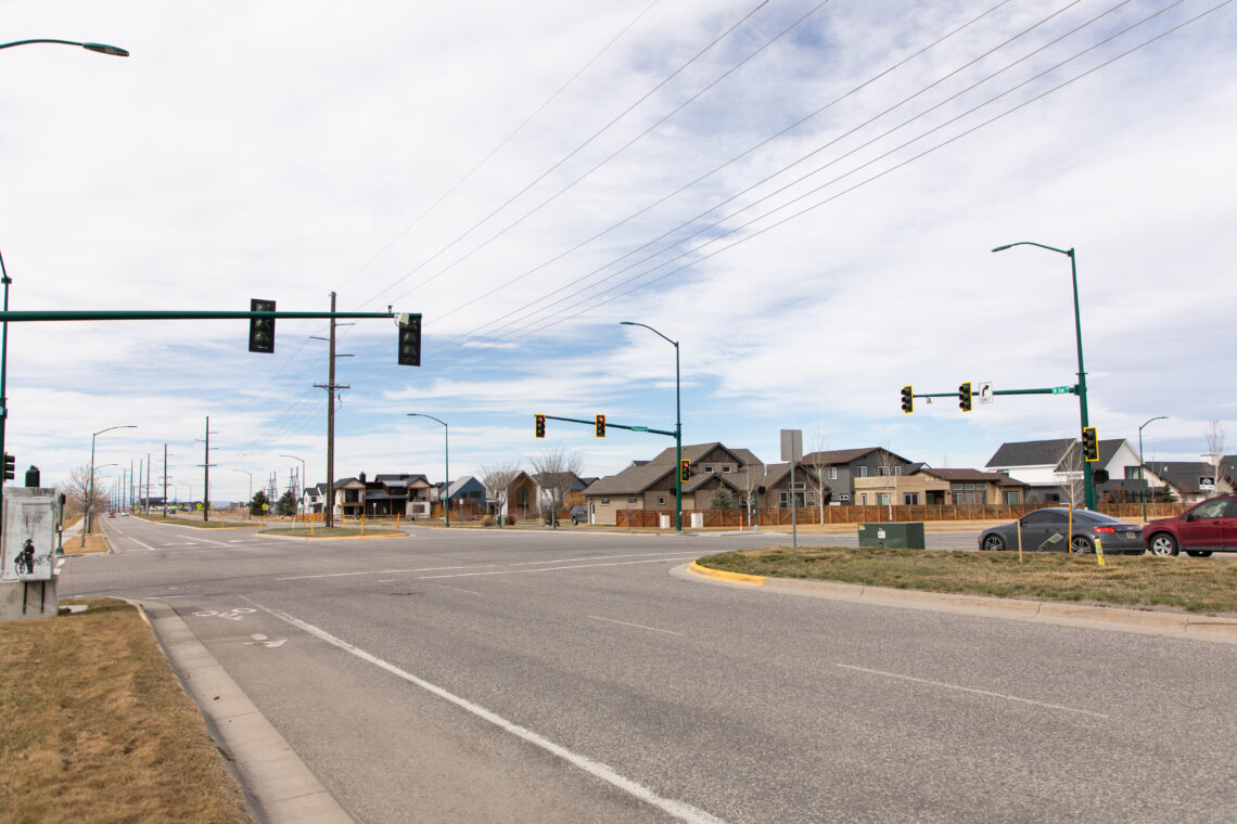 Wide view of a suburban intersection with traffic lights, power lines overhead, and houses in the background under a cloudy sky.