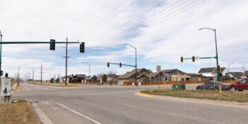 Wide view of a suburban intersection with traffic lights, power lines overhead, and houses in the background under a cloudy sky.