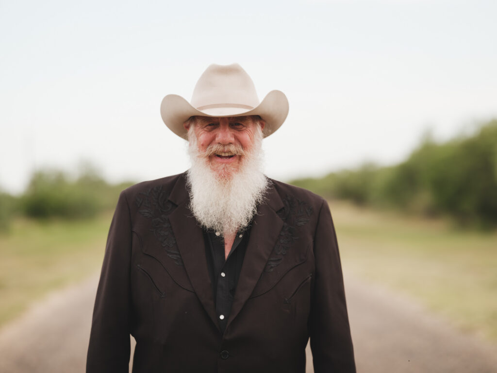 Smiling elderly man with a white beard wearing a light cowboy hat, standing outdoors on a sunny day.