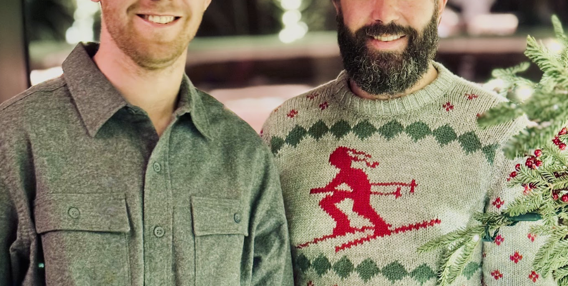 Two men smiling for a photo at a holiday gathering; one in a gray shirt, the other in a red-reindeer sweater.