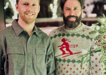 Two men smiling for a photo at a holiday gathering; one in a gray shirt, the other in a red-reindeer sweater.