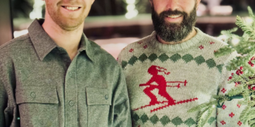 Two men smiling for a photo at a holiday gathering; one in a gray shirt, the other in a red-reindeer sweater.