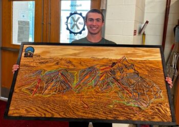 Smiling young man in a green shirt holds a large framed wood artwork depicting a colorful mountain landscape indoors, near a double door.