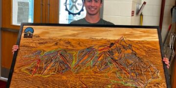 Smiling young man in a green shirt holds a large framed wood artwork depicting a colorful mountain landscape indoors, near a double door.