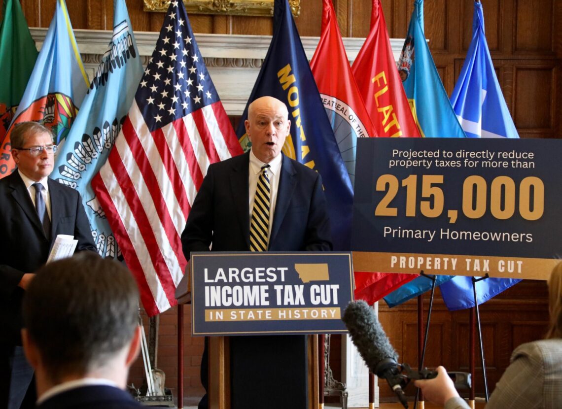 Speaker at podium flanked by multiple flags during a press conference or rally about tax cuts and homeowners, with a large banner in the foreground reading 'Largest income tax cut'.
