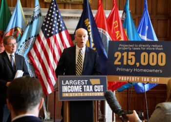 Speaker at podium flanked by multiple flags during a press conference or rally about tax cuts and homeowners, with a large banner in the foreground reading 'Largest income tax cut'.